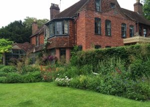 A large brick house with multiple chimneys and a bay window sits nestled in lush, green gardens and colorful flowers, evoking the enchanting charm of a Secret Garden hidden in the Chilterns.