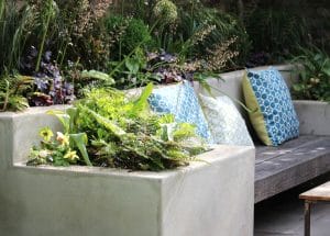 Symmetrical concrete garden bench with blue and green patterned cushions, surrounded by lush green plants in a raised bed.