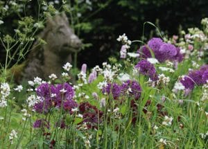 A secret garden with tall grasses and blooming purple and white flowers, where a weathered stone animal sculpture is partially visible in the background, evokes the tranquil beauty of the Chilterns.