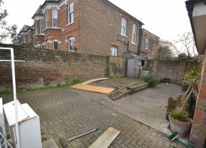 Symmetrical paved backyard with a raised concrete bed, small shed, potted plants, and brick walls; neighboring brick houses are visible in the background.
