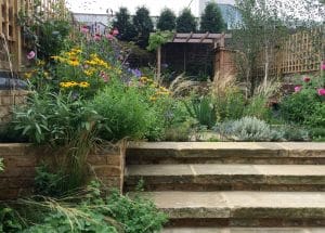 Stone steps lead up to a garden with dense, colorful flowers, tall grasses, shrubs, and a wooden pergola in the background.