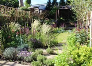 A well-maintained garden with cottage style planting features a variety of flowering plants, tall grasses, shrubs, a stone path, and a wooden pergola in the background.