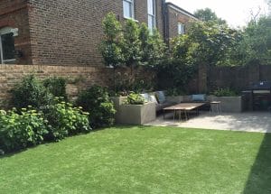 A symmetrical backyard features artificial grass, concrete planters, benches with cushions, a small table, and mature greenery set against a classic brick wall.