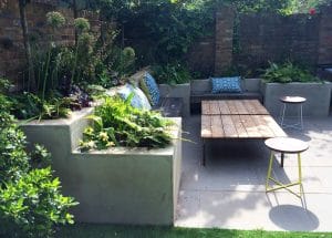 A modern, symmetrical patio features built-in concrete benches, patterned cushions, a wooden table, stools, and lush plantings against a brick wall.
