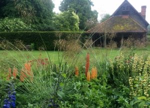 A rustic wooden cottage with a mossy roof sits behind a lush Secret Garden, filled with tall grasses, orange and yellow flowers, and dense greenery—tucked away like a hidden gem in the Chilterns.