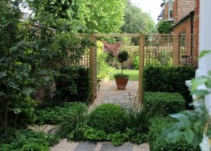 A landscaped garden with symmetrical gravel paths, trimmed bushes, and a potted tree centered under a wooden garden arbor, with a grassy lawn in the background.