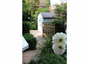 A small, symmetrical blue shed with a gray roof sits behind a wooden gate in a lush garden filled with green bushes, gravel paths, and white flowers in the foreground.