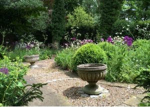 Two stone planters sit on a gravel and stone pathway in a Secret Garden, surrounded by green foliage, bushes, and blooming purple and white flowers reminiscent of the tranquil Chilterns.