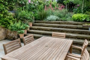 A wooden outdoor dining table with six chairs sits on a patio; stone steps lead up to a garden featuring cottage style planting with various plants and flowers.