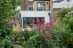 A garden with tall green plants and cottage style planting of pink flowers leads to a patio area outside a modern extension of a brick house with large glass doors.