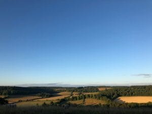 Rolling green and golden fields stretch under a clear blue sky in the Chilterns, with trees and a distant house evoking the charm of a Secret Garden on the horizon.