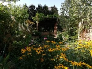 Garden scene with yellow and pink flowers in the foreground, dense green foliage, and a wooden gazebo partially shaded by trees in the background, showcasing charming cottage style planting.