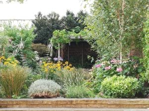 A lush garden with cottage style planting, featuring various flowering plants, shrubs, and a small wooden pergola in the background, bordered by a stone edge.