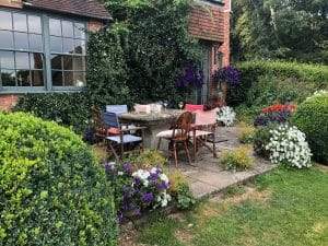 A stone patio with a table and six mismatched chairs is surrounded by flowering plants and shrubs, creating a Secret Garden feel beside a brick house with large windows in the heart of the Chilterns.