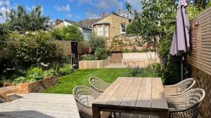 A wooden outdoor dining set sits on a deck overlooking a landscaped garden with grass, brick planters, and oval gardens among the trees, all enclosed by a wooden privacy fence.
