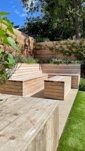 Wooden outdoor benches and table on a stone patio in oval gardens, surrounded by greenery and wooden fencing under a sunny sky.