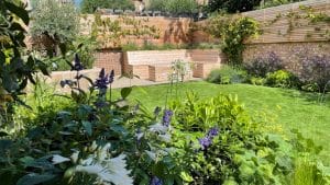 A landscaped garden with green lawn, flower beds, shrubs, and a wooden bench along a brick wall with horizontal wooden fencing—perfect for those who appreciate the charm of oval gardens.