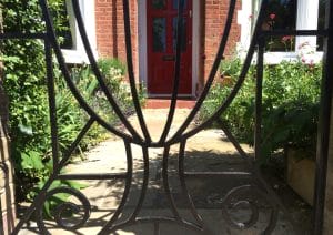 A black metal gate with decorative curves opens onto a stone path through the front garden, leading to a red front door of a brick house, surrounded by green plants and flowers.
