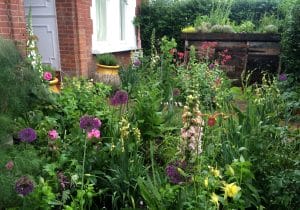 A charming front garden with various flowering plants and lush greenery sits in front of a brick house with a white-framed window and yellow pots. A wooden compost bin is visible in the background.