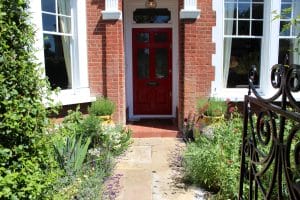 A red front door on a brick house, surrounded by white-framed windows, with a stone path winding through the front garden and plants, leading from a black iron gate.