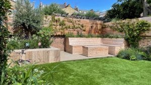 A modern oval garden features a wooden bench with built-in planters, green grass, and various plants along brick and wooden fencing under a clear sky.