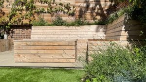 Wooden slatted benches and planter boxes create a raised seating area in landscaped oval gardens, featuring stone paving and lush green grass.