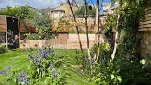 A landscaped oval garden with a wooden fence features raised brick and wood planters, a lush green lawn, and various flowering plants and shrubs.