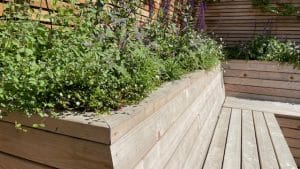 A wooden bench with built-in planters filled with green plants and flowers graces this oval gardens-inspired outdoor space, set against a slatted wooden fence.