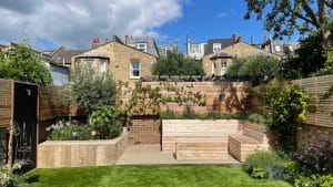 A modern backyard garden with wooden benches, raised planters, a green lawn, and wooden fencing—featuring elegant oval gardens—all set against brick houses under a partly cloudy sky.