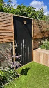 A wooden garden fence with a blackboard panel and copper outdoor light sits amidst greenery in Oval Gardens, featuring a wooden bench on artificial grass for relaxing moments.