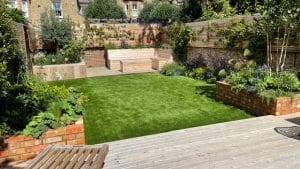 A landscaped backyard with a wooden deck, brick planters filled with greenery, a central grass lawn reminiscent of oval gardens, and wooden fences enclosing the area.