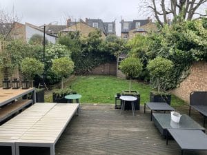 A backyard with a wooden deck, outdoor furniture, potted plants, a grassy lawn with oval gardens, and a shed at the back, surrounded by brick walls and greenery.