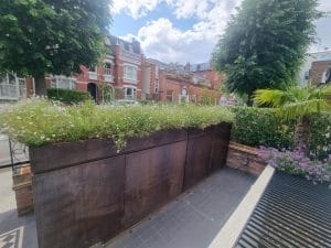 A raised metal planter filled with greenery and flowers sits on a tiled patio in the front garden, with brick houses and trees in the background under a partly cloudy sky.