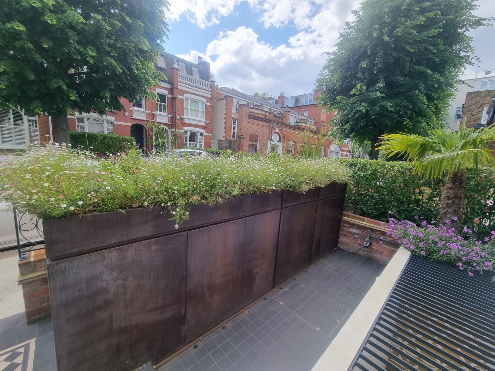 A raised metal planter filled with greenery and flowers sits on a tiled patio in the front garden, with brick houses and trees in the background under a partly cloudy sky.