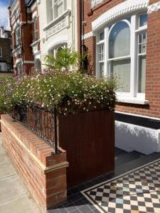 Brick townhouse with white-trimmed windows, a front garden featuring a large planter of daisies, wrought-iron railing, and black-and-white patterned tile by the entrance.