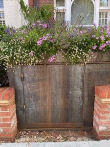 A rusty metal utility box behind a brick wall is topped with a planter of purple and white flowers, adding charm to the front garden in front of a white building with windows.
