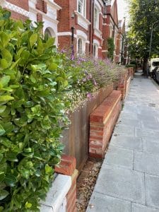 A sidewalk beside brick townhouses features a front garden, with a hedge and flowers in a raised brick and metal planter. Cars are parked along the street.
